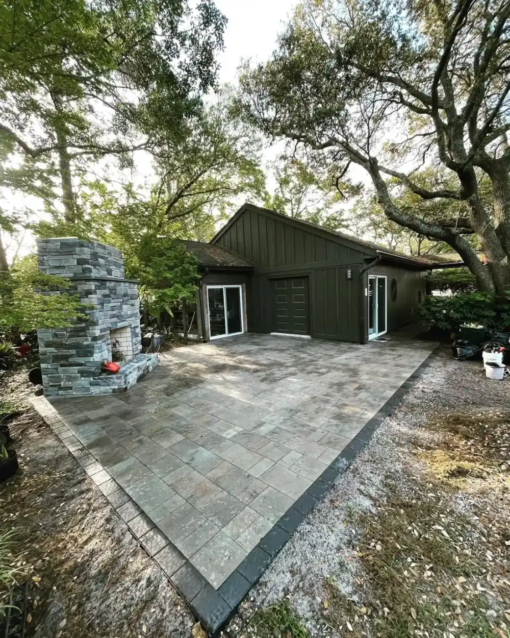 Completed paver terrace and tall stone fireplace beneath mature trees
